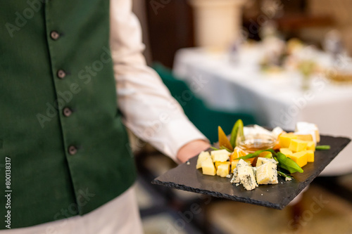 Waiter in green vest and white shirt, presenting a slate tray with assorted gourmet cheeses, embodying fine dining elegance