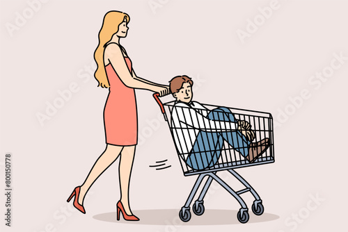 Woman carries man in supermarket carts, for concept of buying boyfriend and financially motivated marriages. Girl gives capricious boyfriend ride on buyer cart because guy doesnt want to go shopping