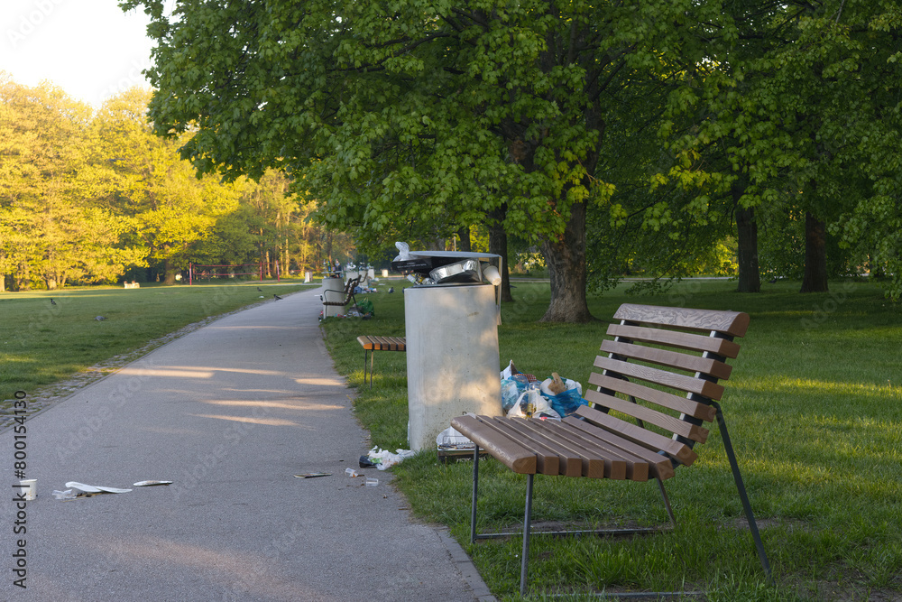 Public trash bin overflowing with plastic bags and snacks wrappings in a green park background on a spring morning day. Public trash bin overflowing with plastic bags and snacks wrappings in a green park background on a spring morning day.
