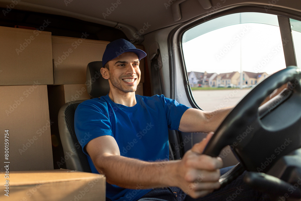 Cheerful man courier in blue uniform on driver's seat of van, delivering parcels, driving car and smiling Cheerful man courier in blue uniform on driver's seat of van, delivering parcels, driving car and smiling