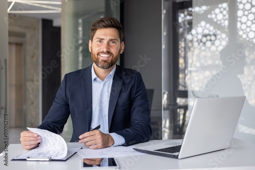 Portrait of a smiling and successful young businessman sitting at a desk with a laptop and signing documents, working with data. Looks confidently into the camera