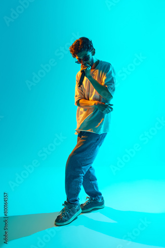 Stylish young man, immersed in music, reading rap, making creative performance against blue, cyan background in neon light. Concept of music, performance, festival, live concert