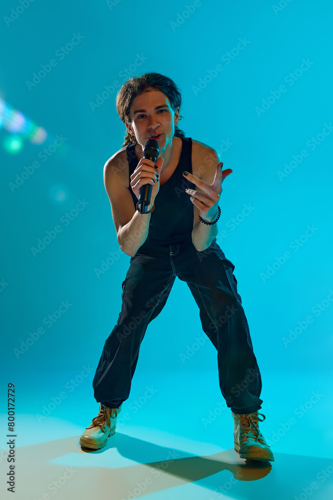 Young man, lost in the rhythm, musician standing on stage and performing against blue, cyan background in neon light. Concept of music, performance, festival, live concert, youth culture Young man, lost in the rhythm, musician standing on stage and performing against blue, cyan background in neon light. Concept of music, performance, festival, live concert, youth culture