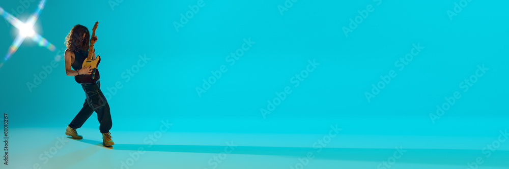 Obraz premium Young guy with log hair playing electric guitar, expressing energy against blue, cyan background in neon. Concept of music, performance, festival, live concert. Banner. Empty space to insert text