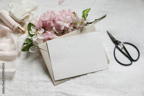 Wedding spring stationery. Feminine scene mockup. Pink blossoming Japanese cherry, apple tree branch. Horizontal blank paper greeting card, black scissors. White linen table background. Flat lay, top