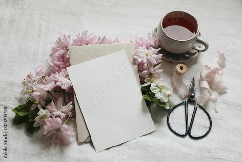Breakfast still life. Spring stationery mockup. Pink blossoming Japanese cherry, apple tree branch. Blank greeting card, invitation. Cup of tea, black scissors. White linen table background. Flatlay