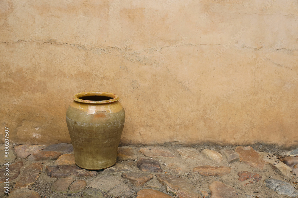Fototapeta premium Rustic yellow clay vase on cobble stone floor, pavement against shabby ochre textured wall, house facade. Old French confit pot or vintage Spanish olive storage pot. Empty copy space, no people.