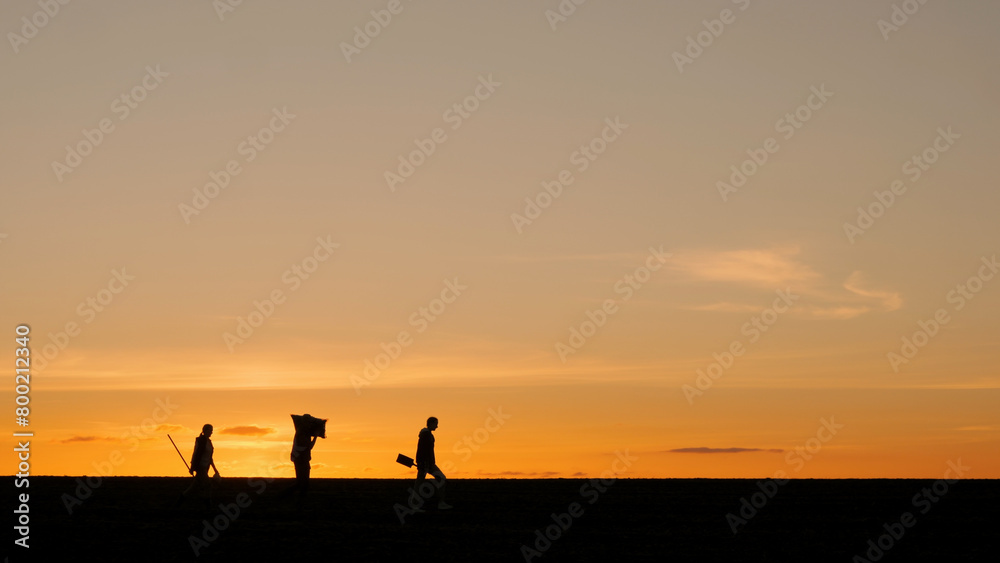 A family of farmers with farming equipment walks through a field at sunset