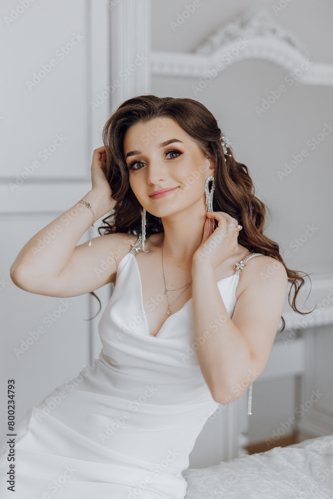 A woman in a white dress is sitting on a bed and putting on her earrings. She has a smile on her face and she is happy A woman in a white dress is sitting on a bed and putting on her earrings. She has a smile on her face and she is happy