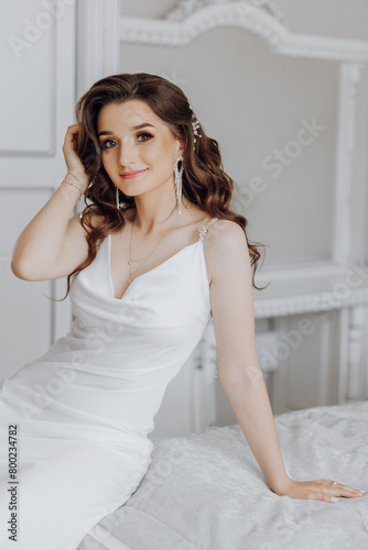 A woman is sitting on a bed wearing a white dress and a necklace. She has long hair and is smiling