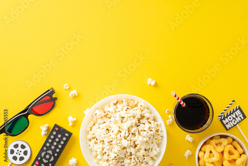 Relaxing cinema experience at home: top view of crunchy popcorn, crisps, soda, 3D glasses. Remote control for online streaming. Movie-inspired decorations on yellow background, space for text or ads