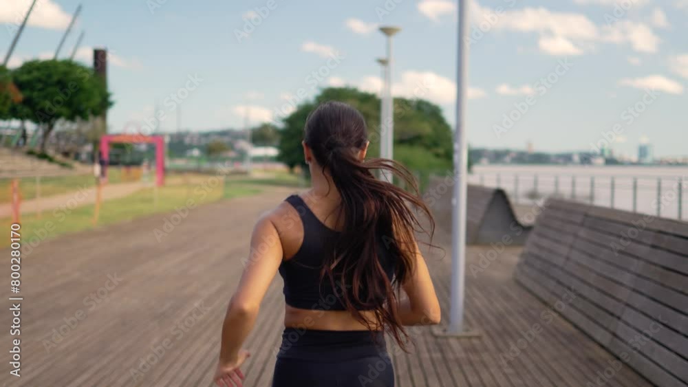 Steadfast jogger advancing in measured strides on a serene riverwalk during a slow-motion workout. Athletic female maintaining her pace with discipline on a tranquil waterfront pathway