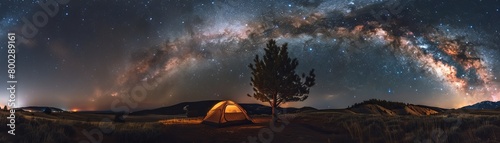 Starry night sky over a lit camping tent and a lone tree in a grassy field