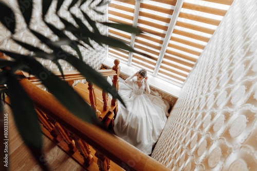 A woman in a white dress is standing on a staircase. The staircase is made of wood and has a leafy plant growing out of it. The woman is looking out of a window, and the room has a warm
