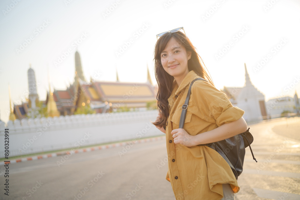 Fototapeta premium Traveler asian woman in her 30s, backpack slung over her shoulder, explores the intricate details of Wat Pra Kaew with childlike wonder. Sunlight dances on the golden rooftops.