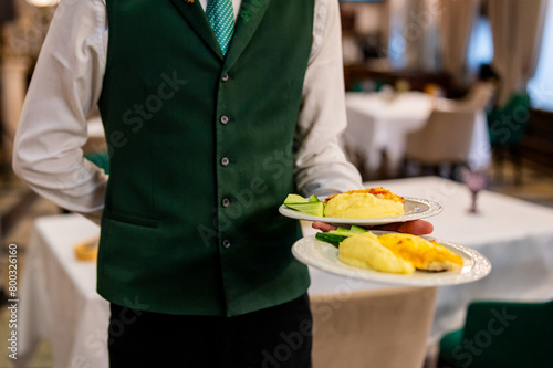 waiter in a green vest and tie serves two plates of delicious food in an elegant dining setting. The professional service of a waiter delivering gourmet meals
