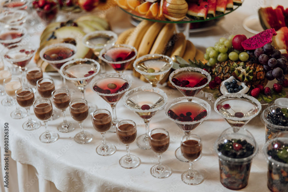 A table full of fruit and drinks, including a variety of cocktails and desserts. Scene is festive and celebratory, as it is a buffet or a party setting