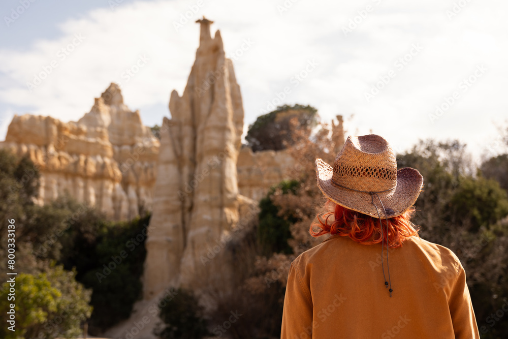 A woman wearing a brown jacket and a hat stands in front of a large rock formation. Concept of adventure and exploration, as the woman is enjoying her time in the desert