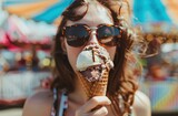a woman with sunglasses licking chocolate and vanilla ice cream in a cone, with a blurry background of a fairground