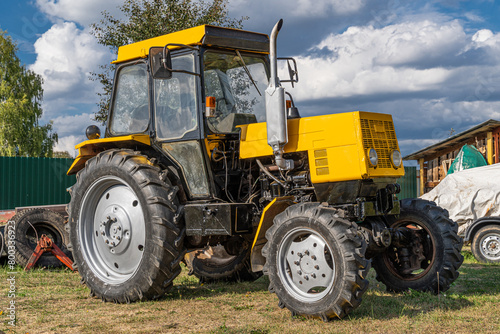 Wheeled Russian diesel tractor of yellow and black color on a sunny day stands on the ground with green grass close-up on the background of a rural yard.
