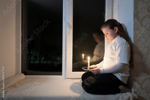 Blackout. A girl in a dark room sits on the windowsill near the window and holds a burning candle in her hands