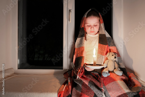 Blackout. A girl, wrapped in a blanket, sits on a windowsill near the window in a dark room and holds a teddy bear and a burning candle in her hands