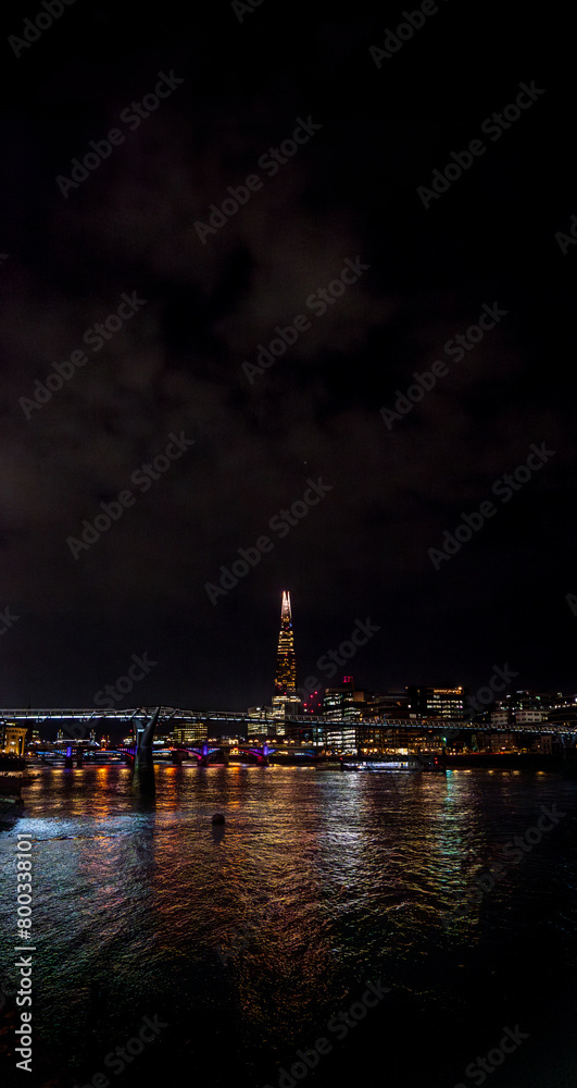 Fototapeta premium The River Thames illuminated by the reflections of the colored lights of the buildings and skyscrapers at night in London with the cloudy night sky with bridges crossing and illuminating the river.
