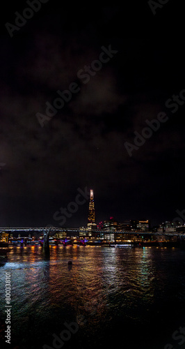 The River Thames illuminated by the reflections of the colored lights of the buildings and skyscrapers at night in London with the cloudy night sky with bridges crossing and illuminating the river.