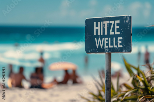 Blue warning sign with German text 'Hitzewelle' (Heat wave) in front of blurry sunny beach with people