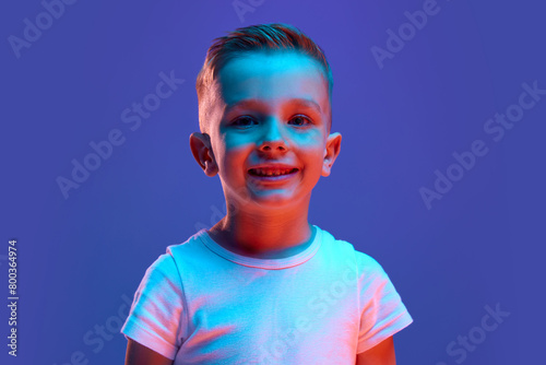 Portrait of charming child, boy dressed white T-shirt looking at camera widely smiling in mixed neon light against blue background. Concept of human emotions, fashion and style, beauty, back to school
