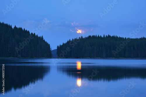 Moon rise up above mountain lake with pine trees.