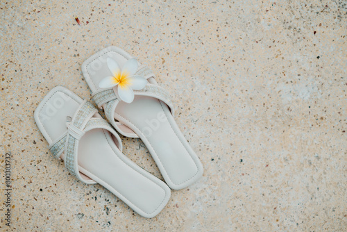 A pair of white flat sandals with a single Frangipani flower on the toe strap placed on the Terrazzo floor