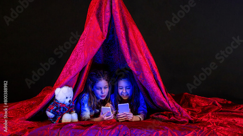 Toddler girls lies on blanket in play tent watching cartoon on tablet with interest, telephone. Little girls in play tent at night using tablet pc under blanket at night. Sisters with telephone