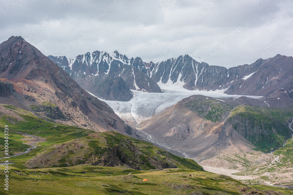 Dramatic landscape with orange tent in alpine valley with view to big glacier tongue and large sharp snow-capped mountain range under gray cloudy sky. Green hills and rocks against ice and sheer crags