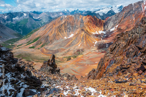 Top view from abyss edge between rocks to multicolor valley with iron river and big sharp rocky ridge of red color. Colorful large mountains in freshly fallen snow in low clouds. Vivid alpine scenery.