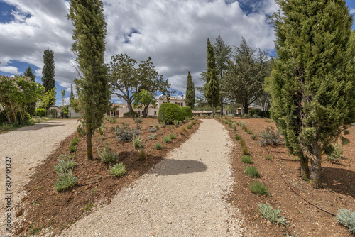 a dirt road in the middle of newly planted gardens with trees and a farmhouse style country house in the background