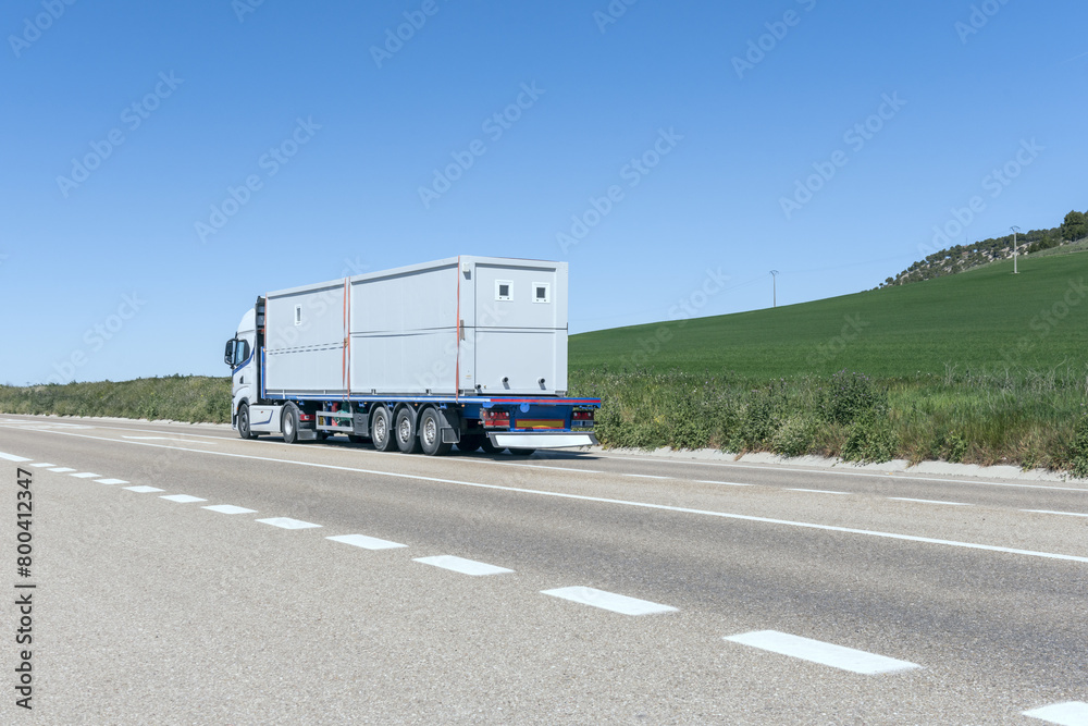 A tractor truck driving with a trailer with some prefabricated construction sheds