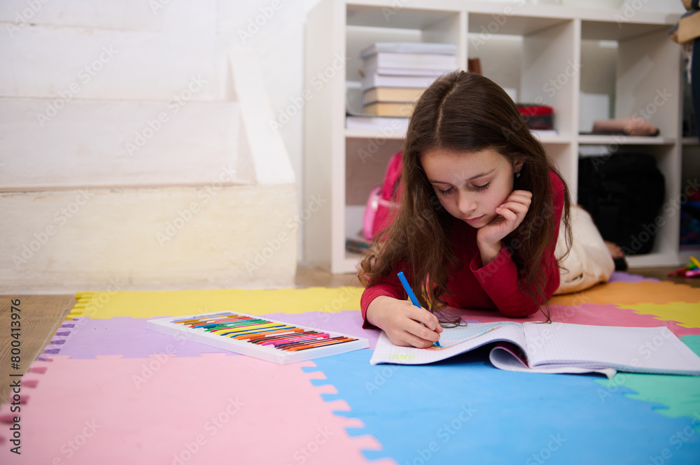 Smart small schoolgirl studying at home, handwriting in notebook. Hardworking focused little girl child drawing picture, doing homework lessons alone, feel motivated at learning, education concept Smart small schoolgirl studying at home, handwriting in notebook. Hardworking focused little girl child drawing picture, doing homework lessons alone, feel motivated at learning, education concept
