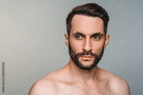 Serious young shirtless man with muscles and beard isolated over grey background. Caucasian sexy shirtless male model guy posing, taking care of his skin hair beard
