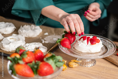 A chef in a green apron assembles strawberry pavlovas in a bright kitchen, with more desserts in soft focus behind. Ideal for culinary websites and dessert blogs.