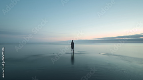 A single person stands in calm, shallow waters under a pastel sky, reflecting on the still surface