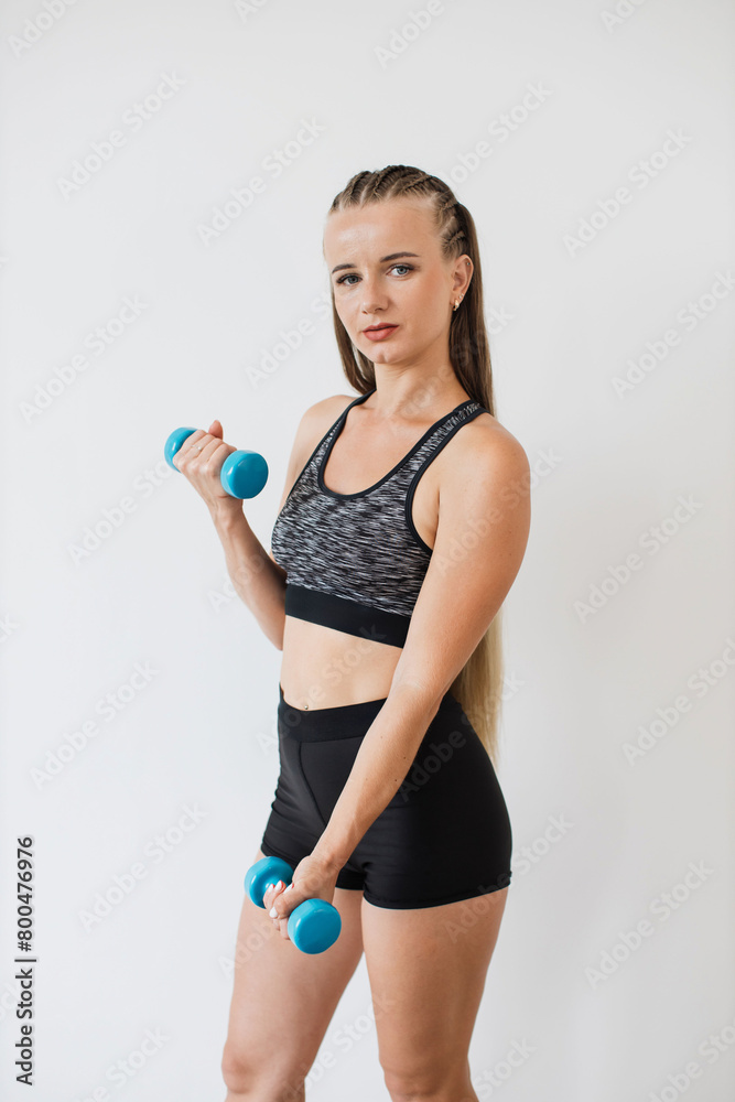 A focused young woman in sports attire performs an exercise routine with blue dumbbells against a white background, exemplifying health and fitness.