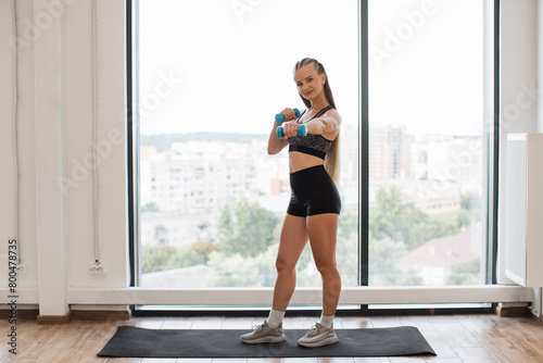 A fit young woman holds blue dumbbells, ready to workout in a bright home environment. She stands on a yoga mat by a large window overlooking the city.