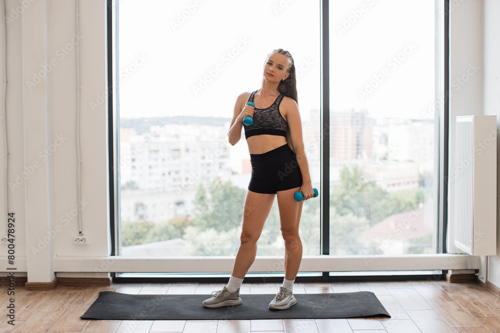 Fit young woman stands on yoga mat by large window, holding blue dumbbells, dressed in sportswear, looks ready for home workout session, illustrating determination, health, and wellness. Fit young woman stands on yoga mat by large window, holding blue dumbbells, dressed in sportswear, looks ready for home workout session, illustrating determination, health, and wellness.