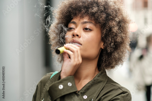 Relaxed curly woman holding an e-cigarette outdoors.