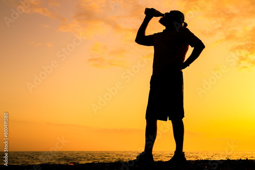 A man is drinking a beverage while standing on a beach at sunset