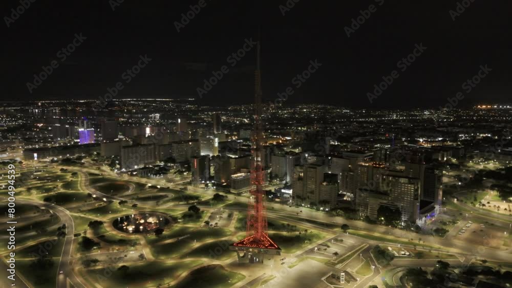 Drone orbits around red TV Tower as it changes to green while overlooking Jardim Burle Marx at night in Brasilia, Brazil