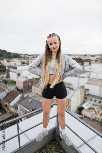 Young woman standing on rooftop with hands on hips confidently overlooking urban landscape. The city's architecture spreads out in all directions, highlighting blend of modern and historic buildings.