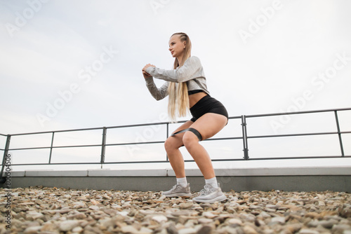 A young athletic woman in sportswear stretches her arms while enjoying an outdoor workout session on a rooftop. Her focused expression and active stance against a cloudy sky backdrop. © sofiko14