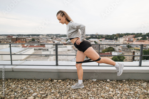 A young woman performs stretching exercises on a rooftop overlooking an urban skyline. She's dressed in sportswear, demonstrating a dynamic stretch, promoting fitness and health. © sofiko14